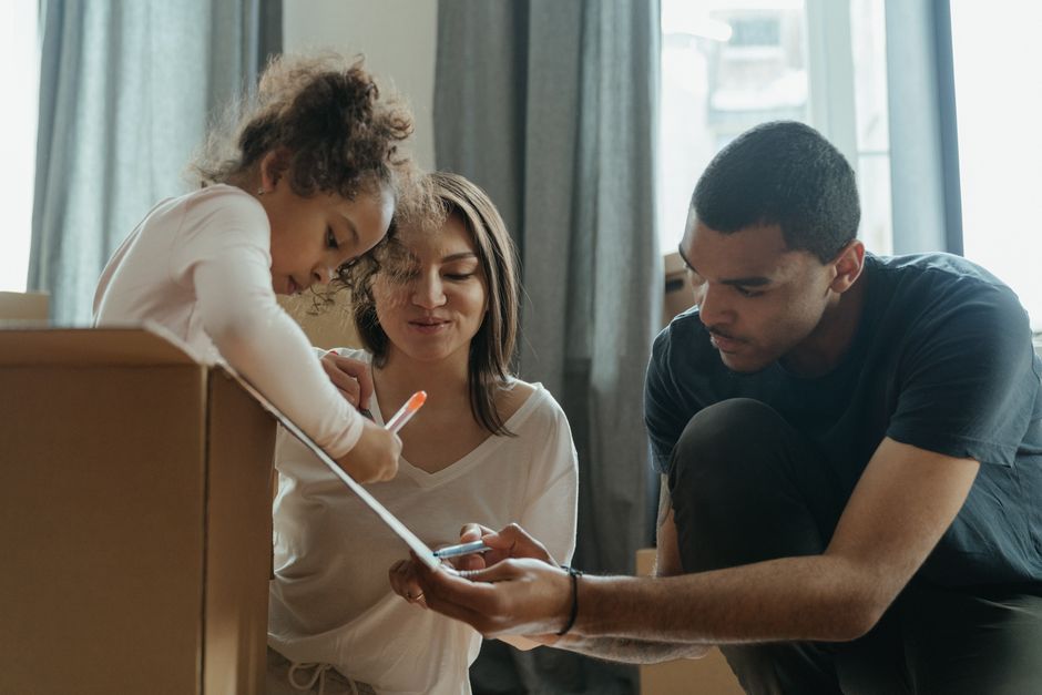 A young girl with curly hair and an adult woman with shoulder-length brown hair are inside a home, inspecting and possibly packing a cardboard box for a home relocation. The woman holds a smartphone in her hand while the girl looks closely at the box. Beside them, a young man with short dark hair, wearing a dark t-shirt, is kneeling and assisting with the packing process, holding a marker or pen. In the background, there are large windows with natural light coming in, and the room features curtains and a neutral colour palette. The scene depicts a collaborative packing effort typical of furniture transport and moving preparations, with the use of cardboard boxes for packing, and the presence of tools or materials used during the packing and loading process, consistent with professional removals services provided by Sutton Man and Van.