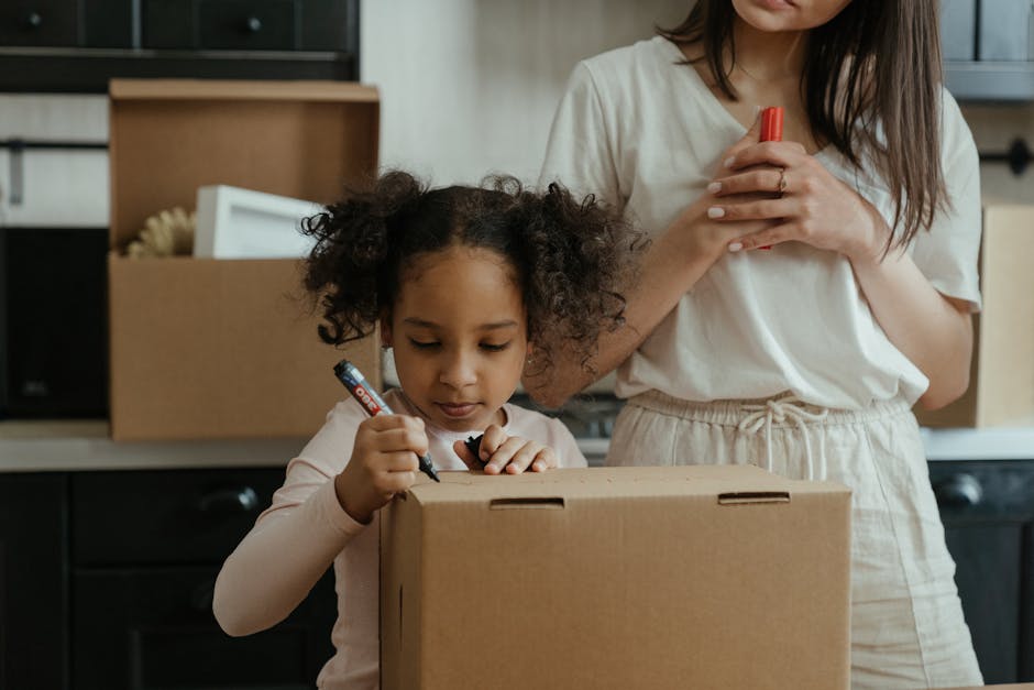 A young girl with curly hair, wearing a light pink long-sleeve top, is seen inside a home, standing at a kitchen counter next to a woman who is partially visible. The girl is holding a black marker and writing on a plain cardboard box, which appears to be part of a home relocation process. Behind them, cardboard boxes and packaging materials are stored on open shelves, and additional boxes are placed on the kitchen counter, indicating packing activity. The woman is wearing a white T-shirt and light-colored trousers, holding a red marker close to her chest while supervising or assisting with the packing task. The environment is well-lit, with natural light coming from an unseen window, and the scene emphasizes careful packing and organisation for a house move, consistent with professional removals and furniture transport services provided by Sutton Man and Van.