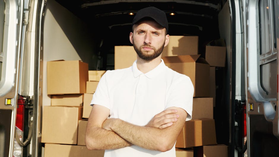 A young man with a beard, wearing a black cap and a white polo shirt, stands inside the back of a moving van with open doors. The van's interior is filled with numerous cardboard boxes of various size