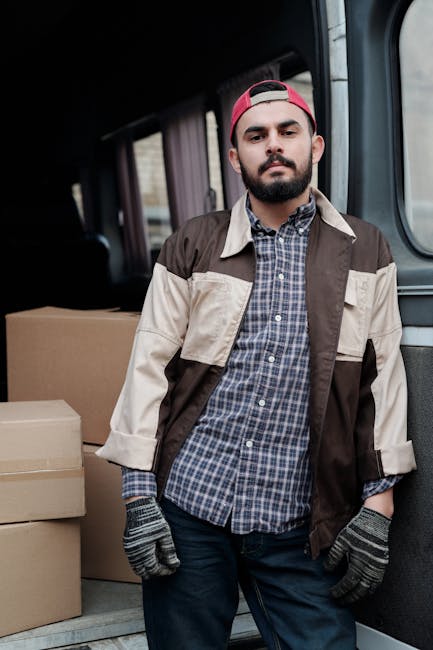A young man with a beard, wearing a black cap and a white polo shirt, stands inside the back of a moving van with open doors. The van's interior is filled with numerous cardboard boxes of various size
