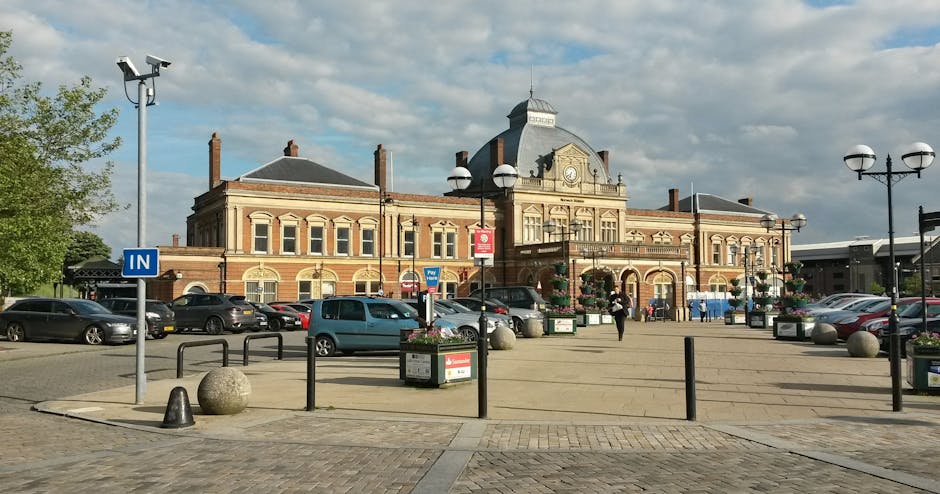 The image depicts the exterior of a historic brick railway station building with a central clock tower and a decorative roof, set against a partly cloudy sky. The station is surrounded by a spacious paved area with parking spaces filled with various cars and a designated loading zone. In the foreground, there are bollards, metal railings, and large spherical concrete planters with flowers, along with streetlamps topped with four round lights, providing illumination for the area. There are also two tall, mounted security cameras positioned at the street corner. Pedestrians are visible walking near the entrance, which is accessed via steps and a ramp, suggesting accessibility considerations. The scene is well-lit by daylight, with some trees visible on the left, adding greenery to the urban setting. This setting aligns with the process of home relocation or furniture transport to or from the station area, fitting with popular local move activities supported by Sutton Man and Van.