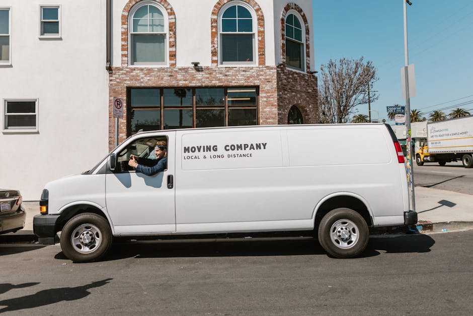 A white moving van parked on a city street in front of a brick building with arched windows during daylight. The van features the company's name, Sutton Man and Van, and the words 'Moving Company, Local & Long Distance' printed on the side. A man is sitting in the driver's seat, visible through the open window, wearing sunglasses and a dark jacket. Visible behind the van are parked cars, including a silver vehicle partially seen at the front, and a yellow truck further down the road. The scene includes a sidewalk with a lamppost and street signs, suggesting an urban environment. The image captures a typical home relocation or furniture transport scenario, with the vehicle ready for loading or unloading inside a busy residential or commercial area, supporting professional moving services provided by Sutton Man and Van.